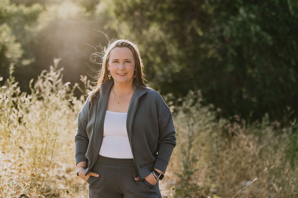 A photo of Rebe standing in a field with her hands in her pockets, smiling directly at the camera.